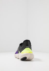 Scarpa sportiva nera, viola e giallo fluorescente con suola bianca curva, mostrata da dietro su una superficie bianca e uno sfondo bianco.