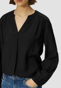 Zwarte lichtgewicht blouse met een V-hals, geplooide details op de schouders en lange mouwen met elastische manchetten, gecombineerd met blauwe jeans.