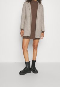 Béžový dlouhý blazer přes hnědé žebrované šaty, doplněný černými platformovými vojenskými botami se zavazováním na tkaničky a texturovanými podrážkami.