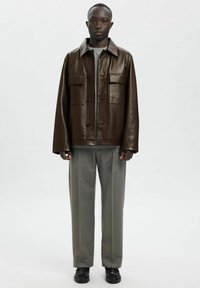 Rjava usnjena jakna z velikimi žepki na prsih, zapenjanjem na gumbe in mehkim ovratnikom; kombinirana s sivimi širokimi hlačami in črnimi čevlji.