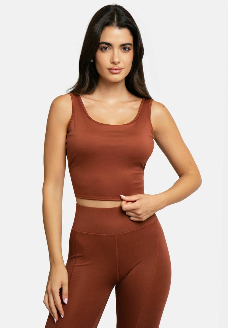 Femme aux longs cheveux foncés portant un débardeur couleur rouille assorti et des leggings taille haute sur un fond clair uni.