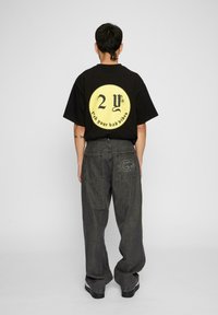 Svart oversized t-skjorte med sirkulært gult grafikk på baksiden, som har stilisert tekst. Kombinert med løse mørke jeans og svarte sko.
