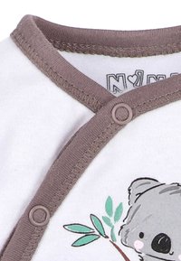Witte onesie met bruin accent, met een illustratie van een koala en groene bladeren. Bevestigd met ronde knopen aan de zijkant. Zachte stofkwaliteit.