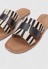 Sandalias planas tipo slide que presentan un patrón de rayas en crema y negro con detalles de cuero. Plantilla suave de color marrón con un acabado liso y diseño abierto.