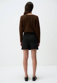 Bruine gebreide trui met geribde textuur, gecombineerd met een zwarte shorts met hoge taille. Witte achtergrond, zichtbare zwarte schoenen.