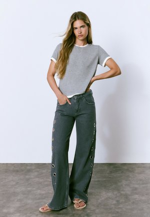 Young woman with long hair wearing gray short-sleeve shirt, wide-leg dark jeans with metal eyelets on sides, and beige sandals, standing indoors.