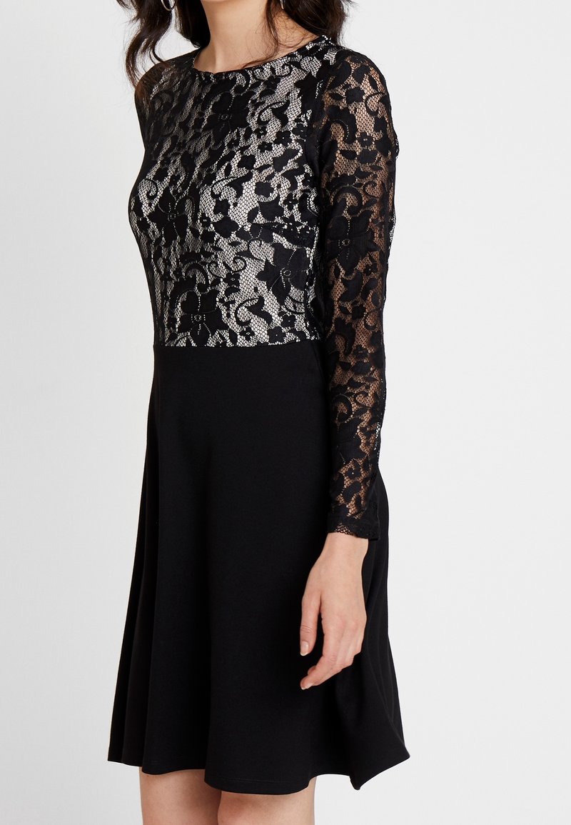 Femme portant une robe noire avec un haut et des manches en dentelle florale, jupe noire unie, debout devant un fond clair uni.