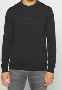 Svart stickad långärmad tröja med rund halsringning. Har tonad broderad text på bröstet. Tyget ser mjukt och slätt ut.