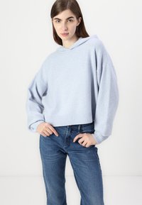 Lys blå, kort sweatshirt med hette, ribbestrikket nederkant og løstsittende ermer, kombinert med klassiske blå jeans. Myk tekstur.