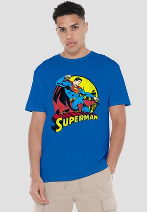 Young man wearing bright blue t-shirt with vintage Superman graphic and beige cargo shorts, standing against plain background.