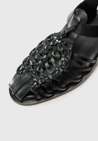 Sandalia plana de piel negra con un diseño trenzado en la parte superior, con acentos suaves y texturizados, punta abierta y forma minimalista.