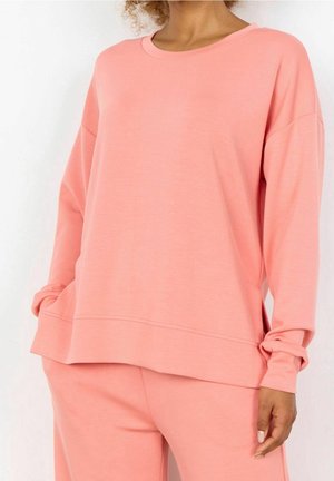 Vrouw met een loszittend roze sweatshirt met lange mouwen en bijpassende roze broek, staand met handen in de zakken tegen een witte achtergrond.