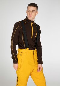 Hnedý zip-up top s rôznymi žltými akcentmi a dlhými rukávmi, v kombinácii so žiarivo žltými snehovými nohavicami, ktoré majú nastaviteľné čierne traky.