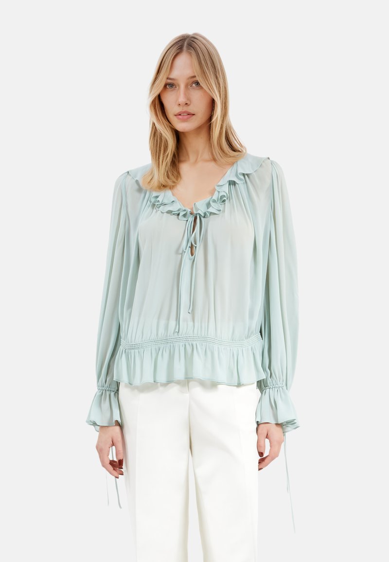 Blusa azzurro chiaro con scollatura arricciata, maniche lunghe con polsini voluminosi, laccio con cordoncino e vita arricciata. Tessuto morbido e fluido.