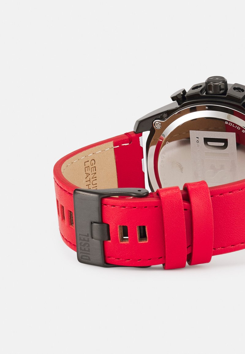 Piros bőrből készült óra szíj fekete fém csattal; vastag, sima textúrával és több állítólyukkal rendelkezik. Az ezüst színű tokon áttetsző hátlap található.