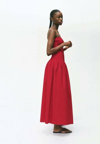 Robe longue sans bretelles rouge avec un corsage smocké et une jupe fluide, assortie à des tongs noires. Texture lisse et design simple.