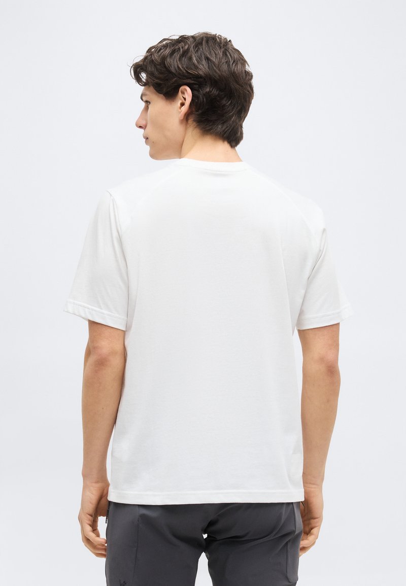 Young man seen from behind wearing a plain white short-sleeve t-shirt and dark gray pants against a light background.