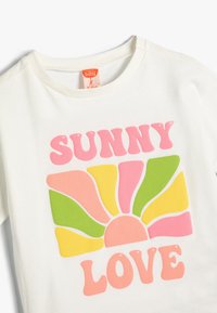 Hvid bomulds t-shirt med et farverigt grafisk motiv med teksten "SUNNY LOVE" i pink, orange, gul, grøn og ferskenfarvet solstrålemønster.