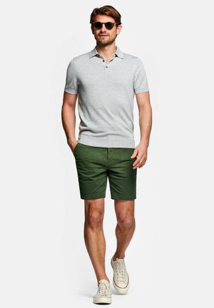Uomo con occhiali da sole, polo grigio chiaro a maniche corte, pantaloncini verdi e scarpe da ginnastica bianche che cammina con una mano in tasca.