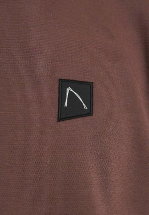 Bruine stof met een gladde textuur, voorzien van een klein zwart rechthoekig patch versierd met een wit abstract ontwerp.