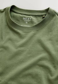 Tričko slim fit v olivovej zelenej farbe, vyrobené z mäkkej bavlny, s okrúhlym výstrihom a minimálnymi švami. Štítok uvádza veľkosť M a pôvod.