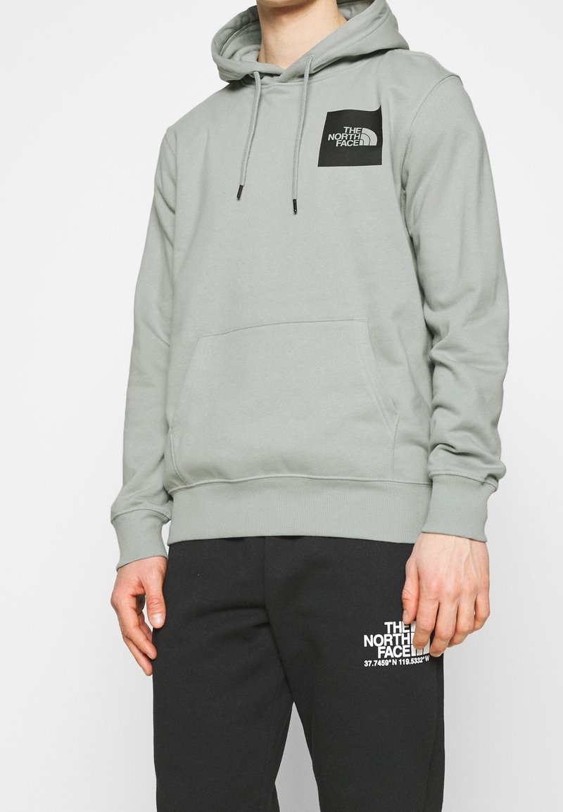Sudadera con capucha de color gris claro con un bolsillo delantero, presenta una etiqueta de logo negra. Combinada con pantalones de chándal negros que tienen coordenadas en blanco.