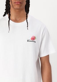 Witte katoenen t-shirt met een kleine geborduurde rode aardbei en de tekst "Bevroren Aardbei" in zwart. Korte mouwen en een ronde hals.