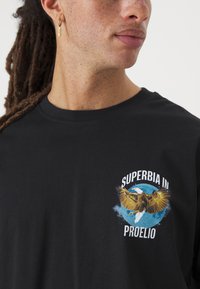 Črna bombažna majica s potiskom orla in belim napisom "SUPERBIA IN PROELIO" nad modrim krožnim dizajnom.