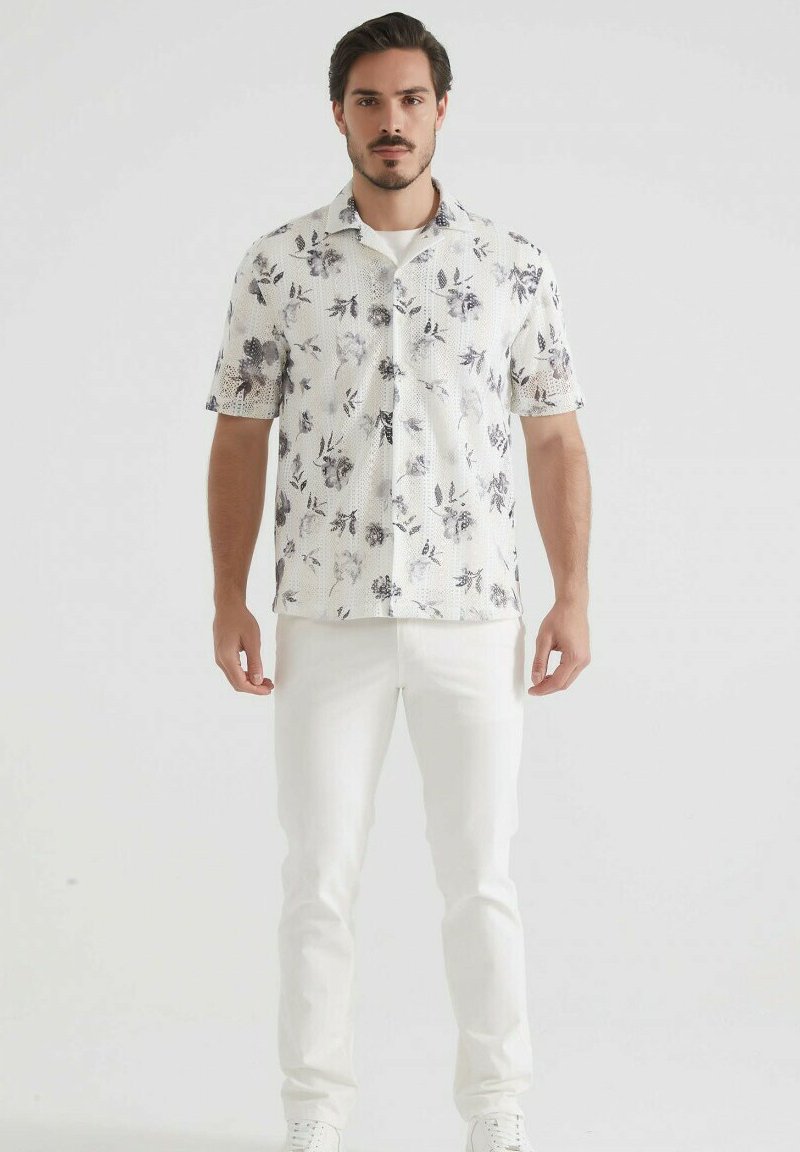 Chemise à manches courtes blanche à motifs floraux avec des accents gris, dotée d'une fermeture à boutons et d'une coupe décontractée, assortie à un pantalon blanc et des baskets.