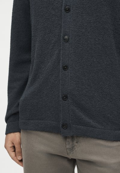 Σκούρα γκρι υφαντή ζακέτα με κουμπιά, φορεμένη πάνω από μπεζ παντελόνια, που δείχνει τα κουμπιά και ένα χέρι που ακουμπάει στο πλάι.