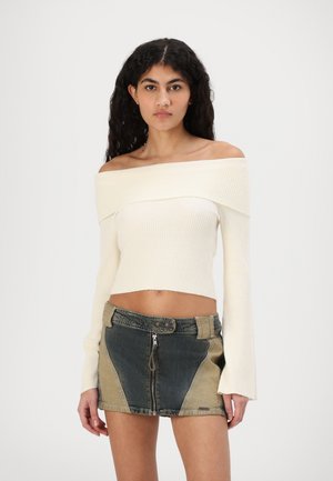 Kreemist ribat täispikkade varrukatega off-the-shoulder pluusist, mis on kombineeritud denimpahkluidega, millel on tõmblukk ja värvilised paneelid.