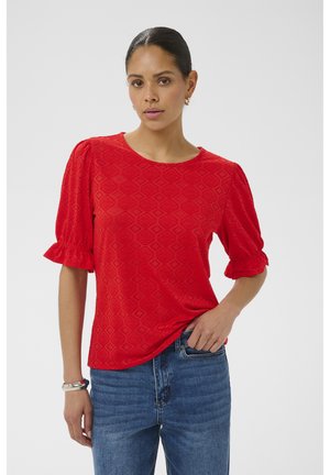 Kobieta w czerwonej bluzce o fakturze z bufiastymi półrękawami i niebieskich dżinsach, stojąca na jednolitym tle.