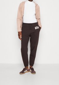 Sudadera de cremallera rosa claro, camiseta de cuello redondo blanca, pantalones de chándal negros con el logo "GAP", zapatillas marrones con rayas blancas y diseño ajustado.