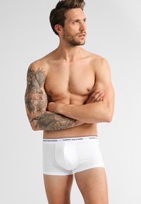 Hvit bomullsboxer med en tett passform, med et logo-belte i marineblått og rødt. Modellen har tatoveringer på armene.