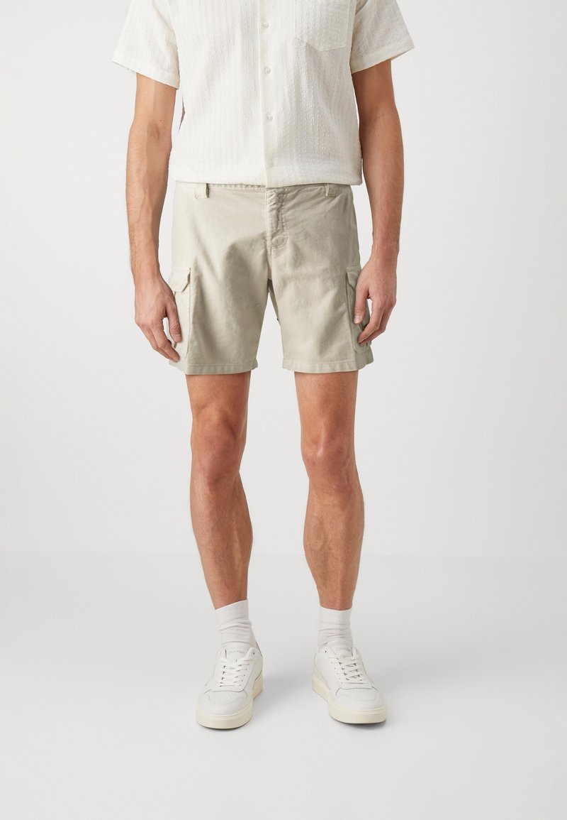 Homme portant un short cargo beige, une chemise blanche à manches courtes avec des boutons, des chaussettes blanches et des baskets blanches, debout devant un fond uni.
