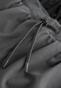 Sort stof med en glat tekstur, der har en justerbar snor med en knyttet ende. Detaljer med samlet materiale tilføjer subtil struktur.