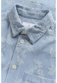 Camicia in denim azzurro chiaro con motivo floreale, colletto a punta, bottoni bianchi e taschino sul petto. Tessuto leggero con una sottile texture.
