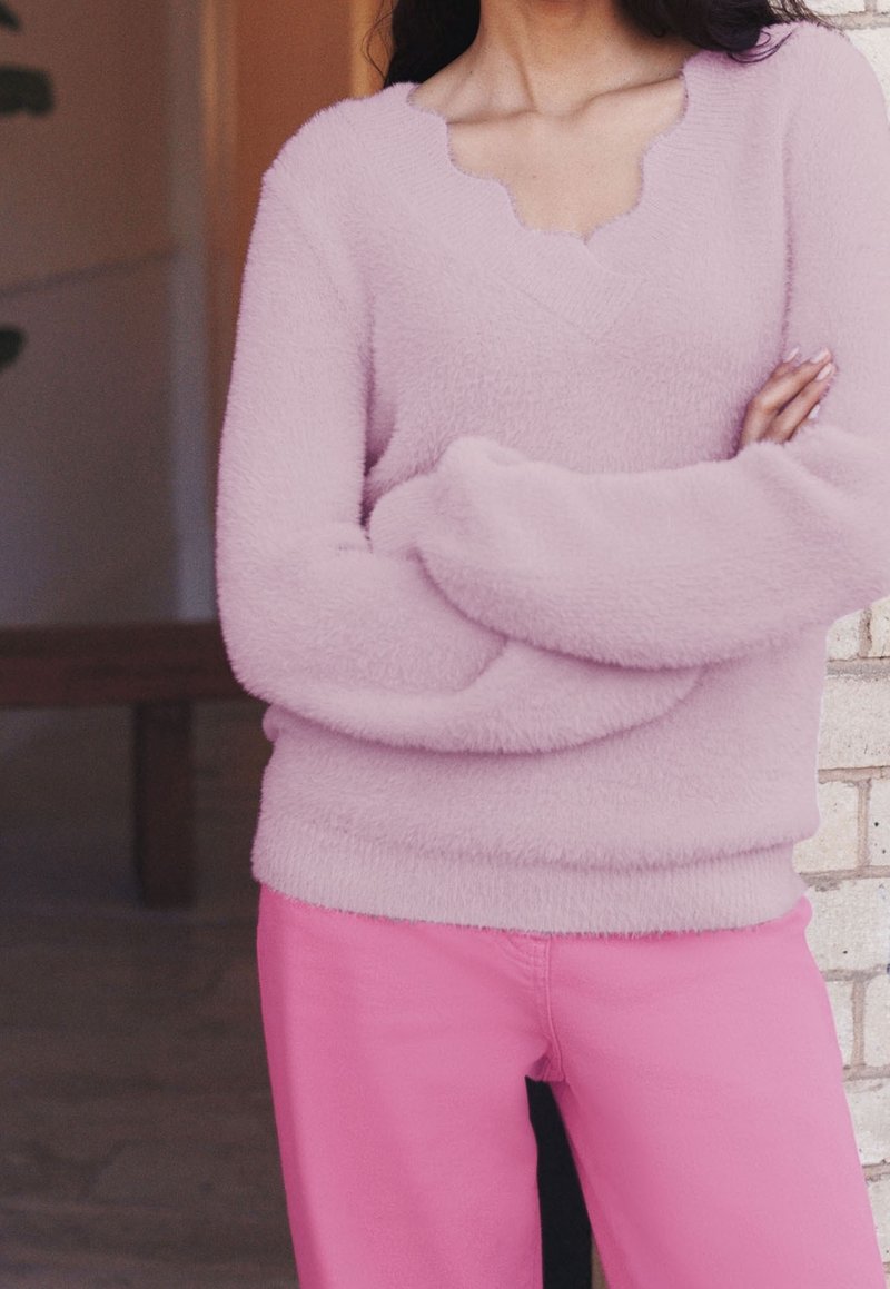 Personne portant un pull duveteux rose clair avec un col festonné et un pantalon rose vif, bras croisés, debout près d'un mur en briques.