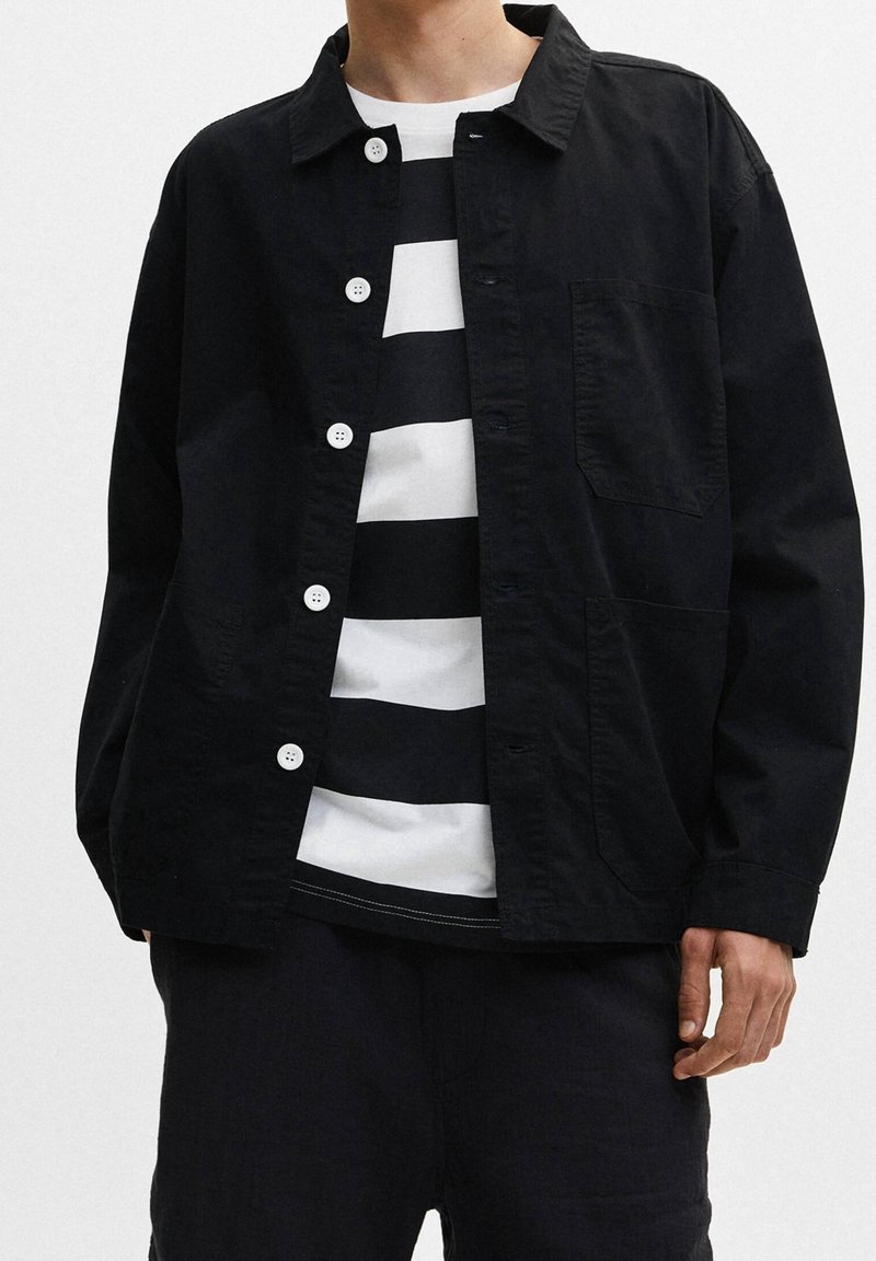 Schwarze leichte Jacke mit Kragen, Knöpfen vorne und einer aufgesetzten Tasche. Trägt man über einem schwarz-weißen gestreiften Hemd.