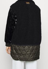 Person som bär en svart texturerad fleece- och quiltad jacka med en G-Star-märkning på övre ryggen, i kombination med vita byxor.