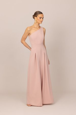 Žena v púdrovo ružovom overale s jedným ramenom a širokými nohavicami, s výraznými zlatými náušnicami, stojaca pred jednoduchým neutrálnym pozadím.