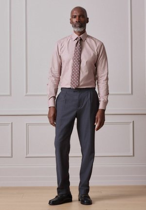 Man with gray beard wearing a pink dress shirt, patterned tie, gray dress pants, and black loafers stands in a room with white paneled walls and wooden floor.