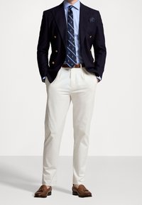Marineblauwe dubbelgeknoopte blazer, lichtblauw overhemd, dasspeld met ankerpatroon, crèmekleurige broek, bruine loafers, wit pochet detail.