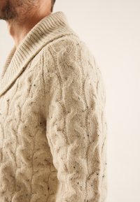 Cremefarbenes Strickpullover mit einem groben Kabelmuster, geripptem Schalkragen und subtilen strukturierten Sprenkeln im gesamten Stoff.