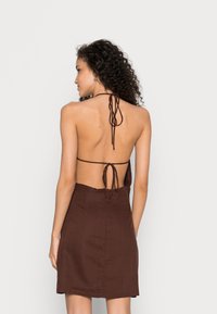 Vestido castanho com design costas nuas, apresentando laços finos e um tecido texturizado. Silhueta simples e reta que termina acima do joelho.