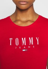 Czerwona bawełniana koszulka z okrągłym dekoltem, z logiem "TOMMY JEANS" w białym kolorze oraz małą niebiesko-białą grafiką flagi. Gładka tekstura.