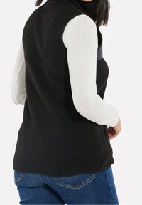 Chaleco de forro polar negro con cuello alto y una tela lisa en contraste en la parte delantera, combinado con una blusa blanca de manga larga y jeans azules.