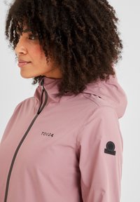 Light pink waterproof jacket with a hood, featuring a front zipper, black logo on the chest and a round patch on the sleeve. Smooth texture.