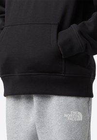 Črna jopa s kapuco z žepom spredaj, rebrastimi manšetami in spodnjim robom; kombinirana s svetlo sivimi trenirkami z logotipom na stegnih.