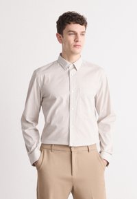 Striped button-up shirt in cream and beige, featuring a collar, long sleeves, and a chest pocket. Paired with beige trousers.